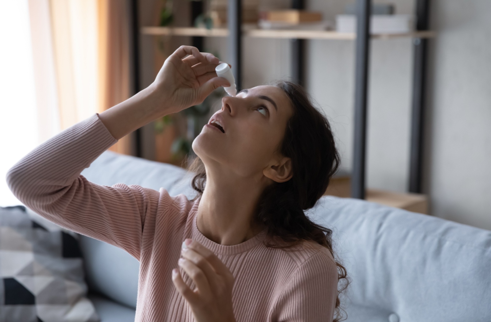 A woman places eye drops in her eyes to get relief from the discomfort she experiences when she blinks.
