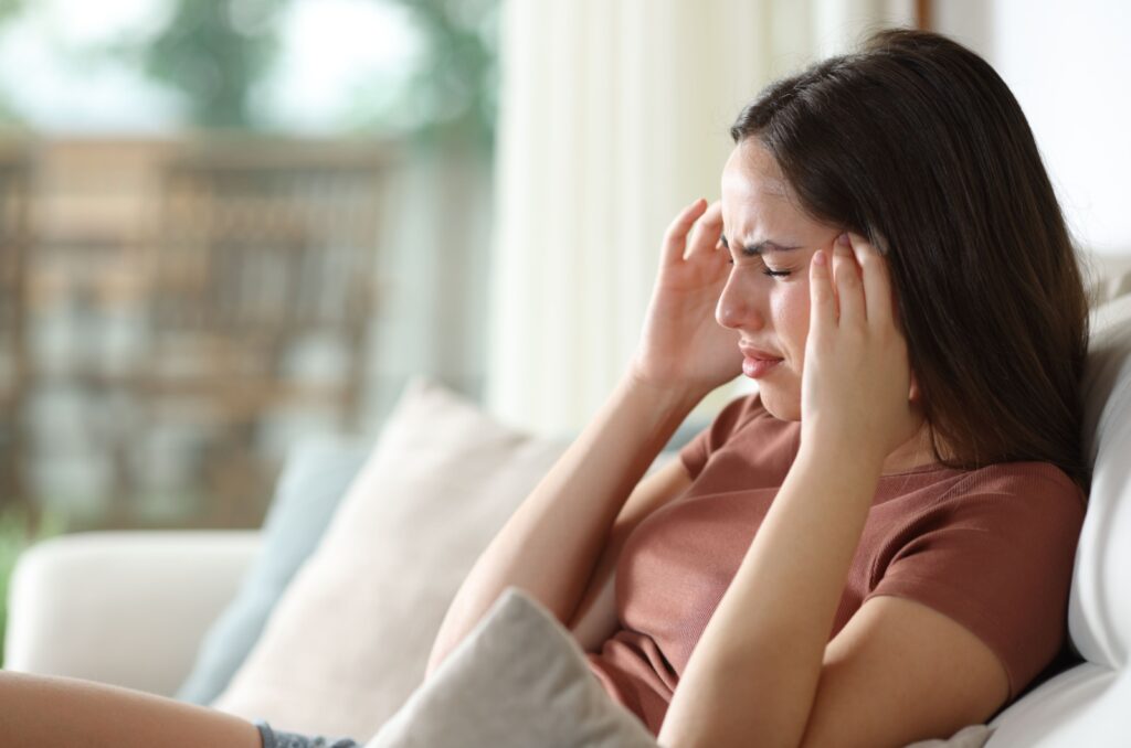 A person with a migraine sitting on a couch with their eyes closed and hands to their temples from light sensitivity.