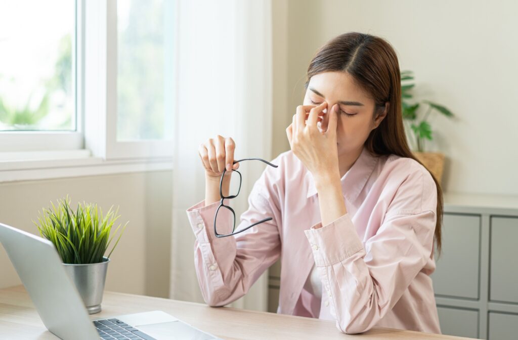 A person holding their glasses in one hand as they rub their eyes due to dry eyes and a headache.